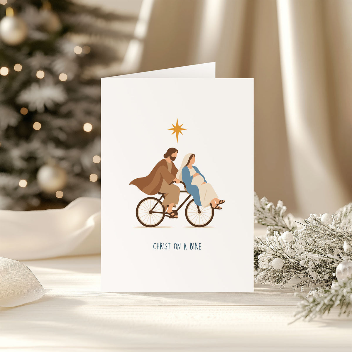 A white greeting card featuring an illustration of Jesus and pregnant Mary riding a bicycle under a gold star, with the text 'CHRIST ON A BIKE' at the bottom. The card is displayed on a light surface with a blurred Christmas tree and holiday decorations in the background.