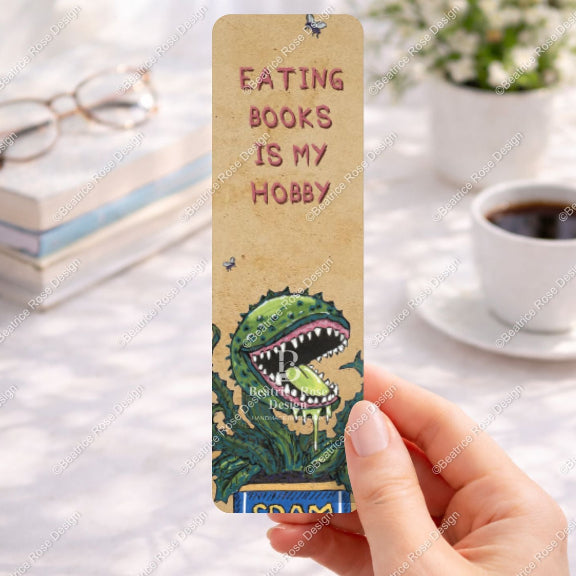 Bookmark with dinosaur illustration and text 'Eating books is my hobby' held by a hand on a desk with books and coffee.