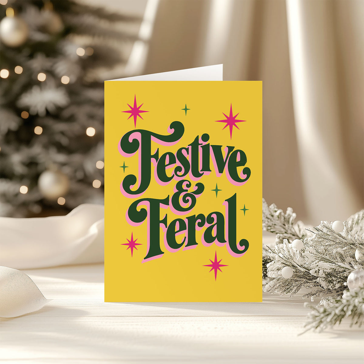 A yellow greeting card with the words 'Festive & Feral' in dark green retro lettering outlined in pink, surrounded by pink and green starburst accents.