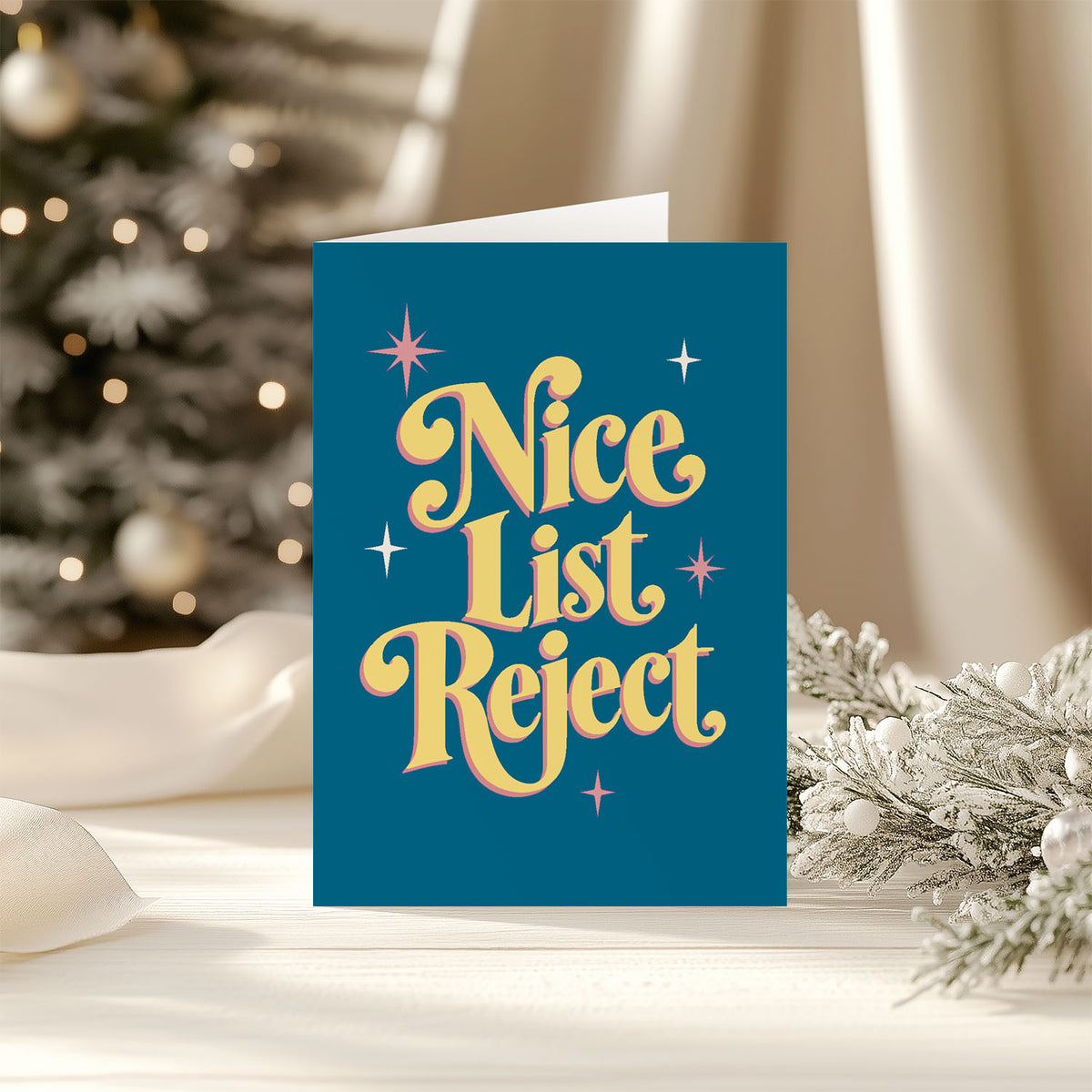 A festive holiday card with the text 'Nice List Reject' in a retro yellow font on a dark teal background, styled on a table with a blurred Christmas tree and holiday decorations in the background.