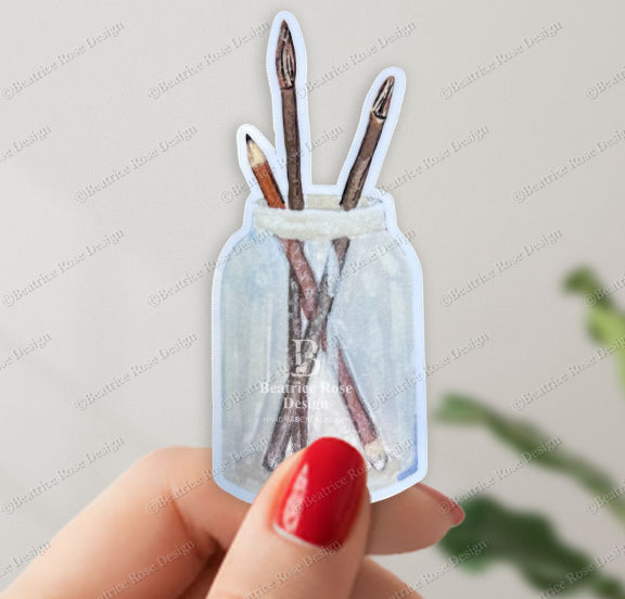 Hand holding a clear sticker with a design of sticks in a bottle against a light background