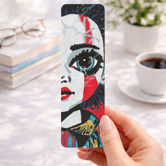 Bookmark with artistic design held by a hand on a desk with coffee and books.