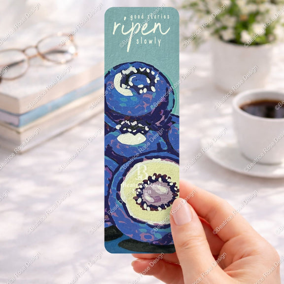 Hand holding a colorful kiwi fruit bookmark with a blurred background of a table with books and coffee.