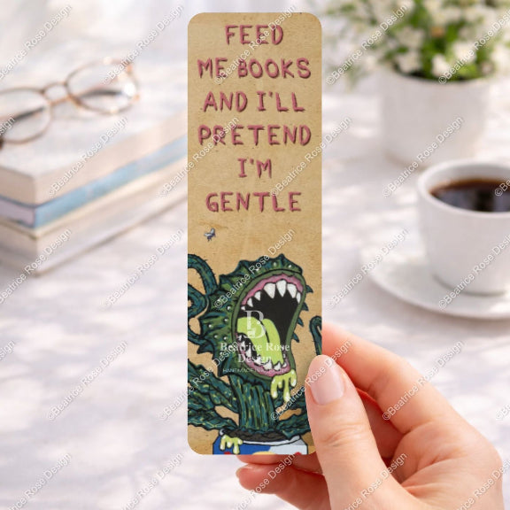 Bookmark with a dragon illustration and text held by a hand on a desk with books and coffee.