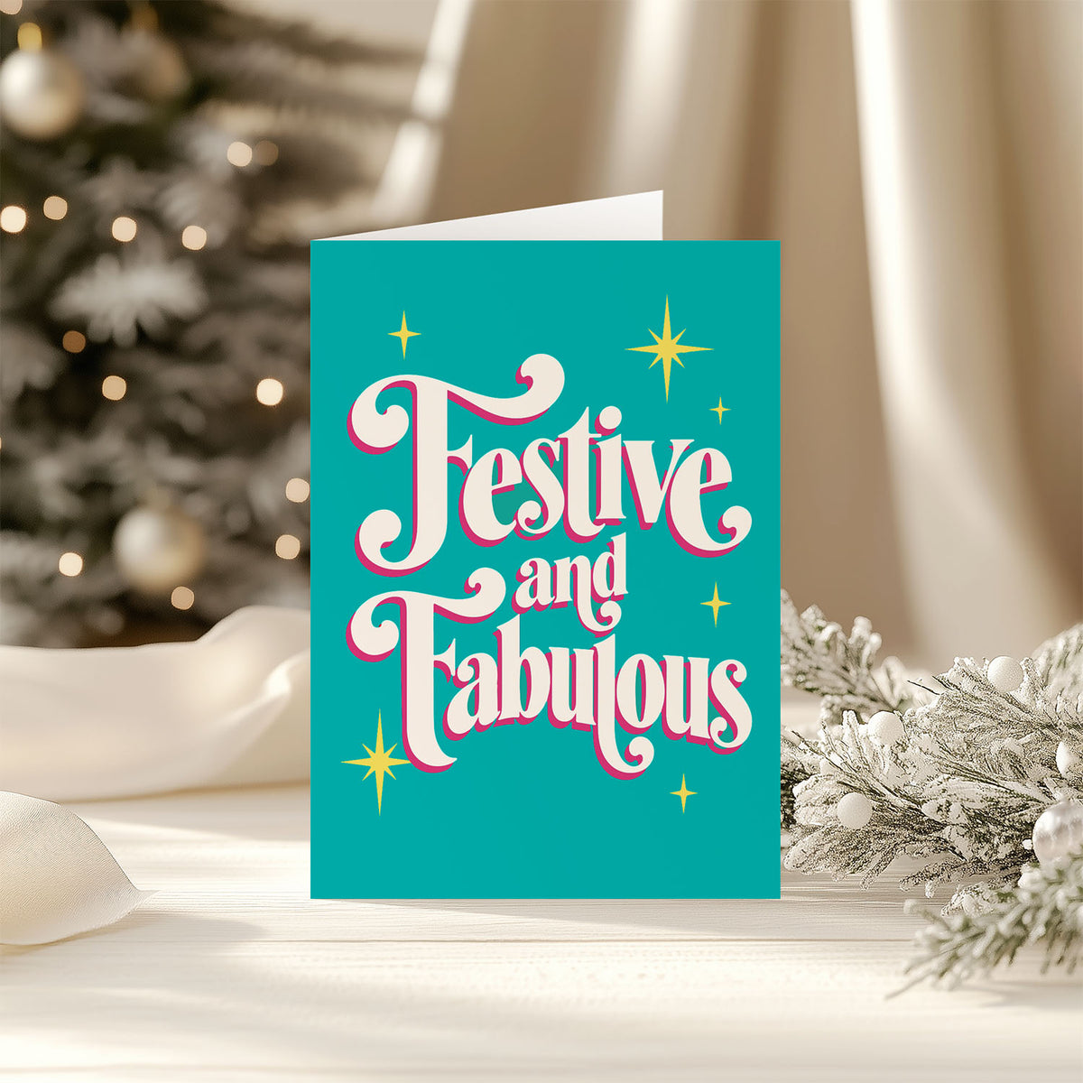 A festive Christmas greeting card with the words 'Festive and Fabulous' in white cursive lettering outlined in pink, set against a bright teal background with gold star accents. The card is displayed on a white surface with a blurred Christmas tree and holiday decorations in the background.