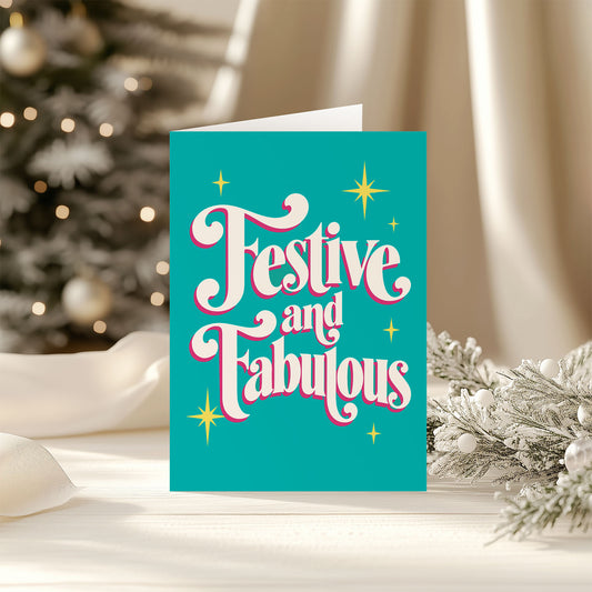 A festive Christmas greeting card with the words 'Festive and Fabulous' in white cursive lettering outlined in pink, set against a bright teal background with gold star accents. The card is displayed on a white surface with a blurred Christmas tree and holiday decorations in the background.