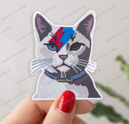 Sticker of a cat with a unique design held by a hand with red nail polish on a light gray background.