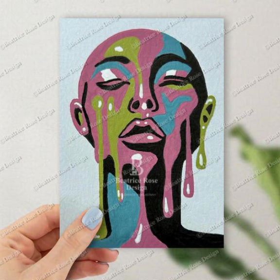 Hand holding a colorful abstract art print of a face with dripping paint on a light blue background.