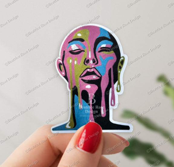Colorful abstract face sticker held by a hand with red nail polish against a light gray background.