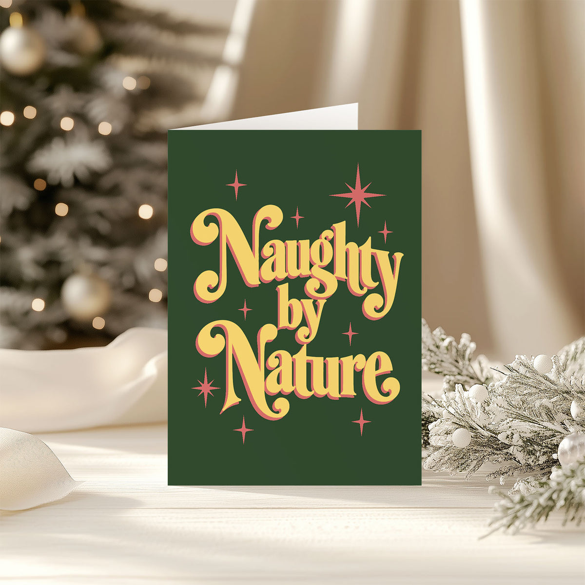 A dark green Christmas greeting card with retro-style yellow and orange lettering that reads 'Naughty by Nature,' styled on a tabletop with a blurred Christmas tree and holiday decorations in the background.