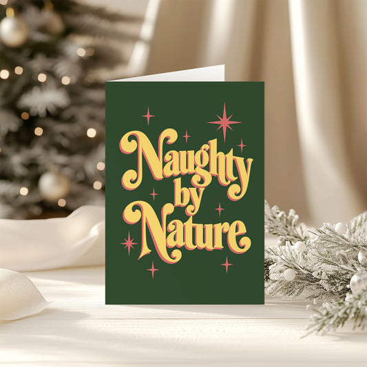 A dark green Christmas greeting card with retro-style yellow and orange lettering that reads 'Naughty by Nature,' styled on a tabletop with a blurred Christmas tree and holiday decorations in the background.
