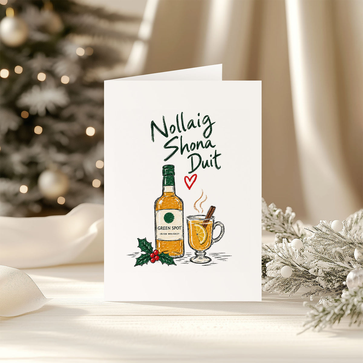 A white greeting card standing upright on a tabletop, printed with the Gaelic phrase 'Nollaig Shona Duit' above an illustration of a Green Spot single malt whiskey bottle and a hot drink with a cinnamon stick. The card is photographed in a warm holiday setting with a blurred Christmas tree, ribbon, and flocked branches in the background.