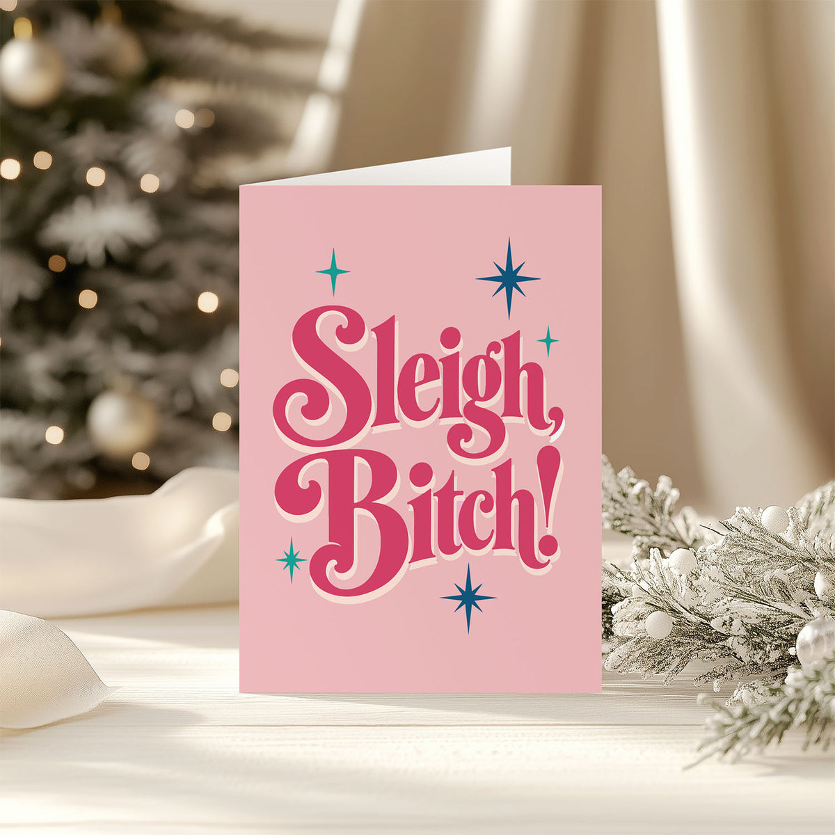 A pink greeting card with the text 'Sleigh, Bitch!' in stylized pink lettering and blue star accents, standing on a tabletop with a blurred Christmas tree and holiday decor in the background.