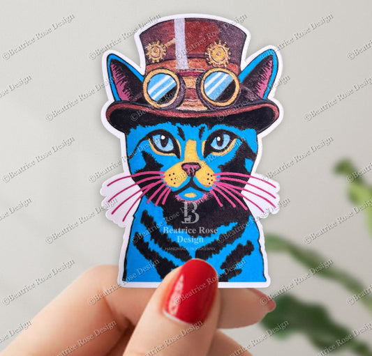 Sticker of a blue cat with goggles and a top hat held by a hand with red nail polish.