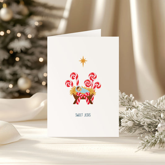A white greeting card standing upright on a tabletop, printed with a nativity scene featuring red-and-white striped candy canes, a gold star, and the words 'SWEET JESUS.' The card is styled in a warm holiday setting with a blurred Christmas tree and white drapery in the background.