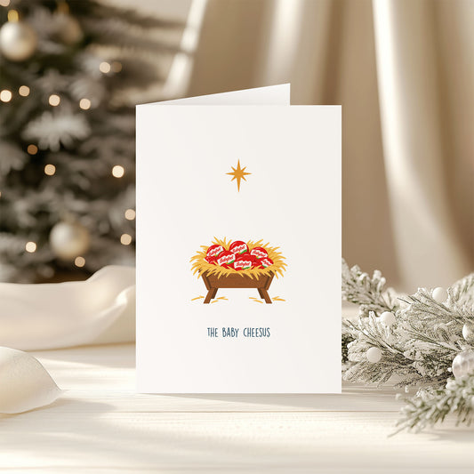 A white greeting card standing upright on a tabletop, printed with a nativity scene featuring a golden star above a manger holding six Babybel cheeses, with the text 'THE BABY CHEESEUS' below. The card is photographed in a softly lit holiday setting with a blurred Christmas tree and white drapery in the background.