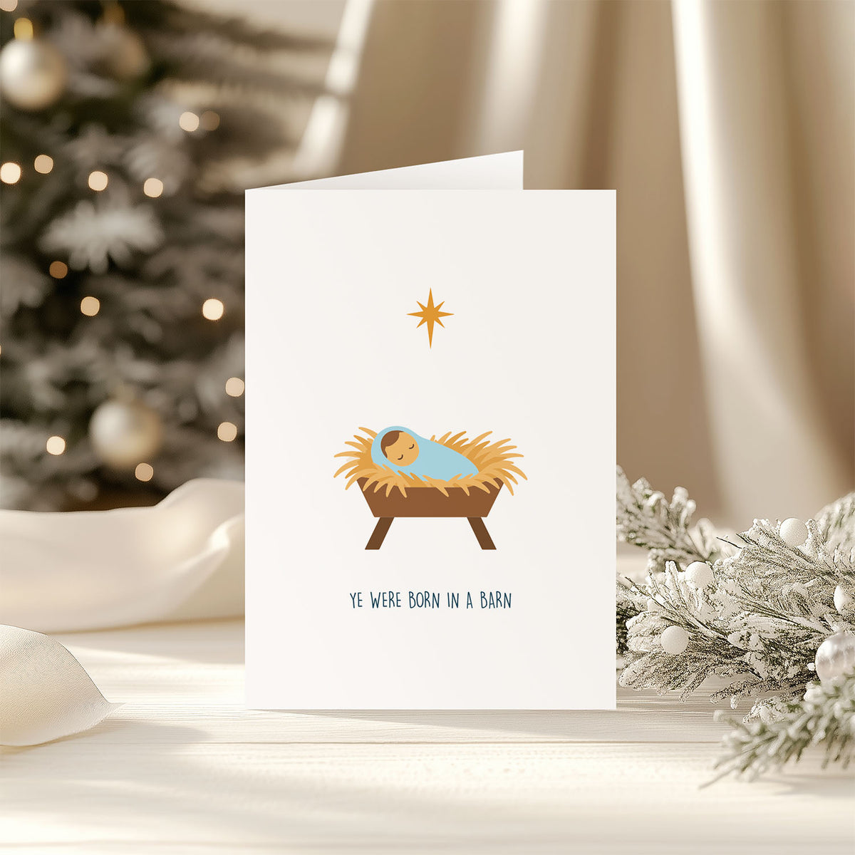 A white greeting card with a simple nativity illustration showing a baby in a blue swaddle in a brown manger, with a gold star above and the text 'YE WERE BORN IN A BARN' below. The card is photographed on a light surface with a blurred Christmas tree and white drapery in the background.