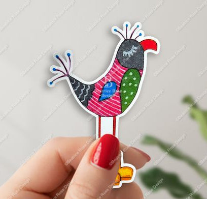 Colorful sticker of an exotic bird held by a hand with red nail polish on a light gray background.