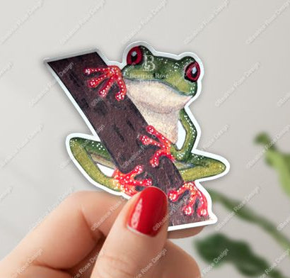 Hand holding a sticker of a frog with red accents on a light gray background