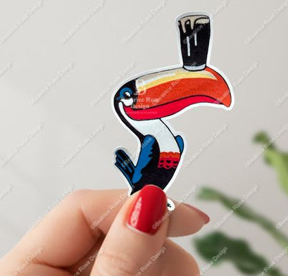 Colorful toucan-shaped sticker held by a hand with red nail polish on a light gray background.